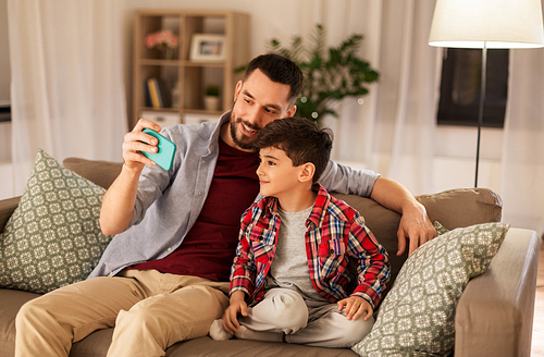 people, family and technology concept - happy father and little son taking selfie by smartphone sitting on sofa at home