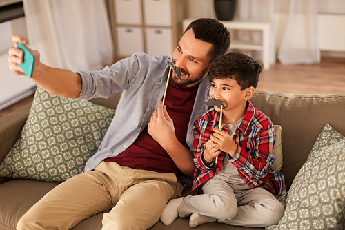 people, family and technology concept - happy father and little son with mustaches party props taking selfie by smartphone sitting on sofa at home