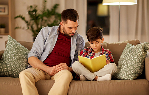 family, childhood, fatherhood, leisure and people concept - father and little son reading book on sofa at home in evening