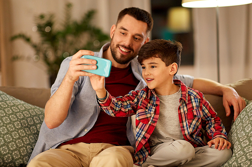 people, family and technology concept - happy father and little son taking selfie by smartphone sitting on sofa at home in evening