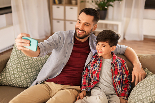 people, family and technology concept - happy father and little son taking selfie by smartphone sitting on sofa at home