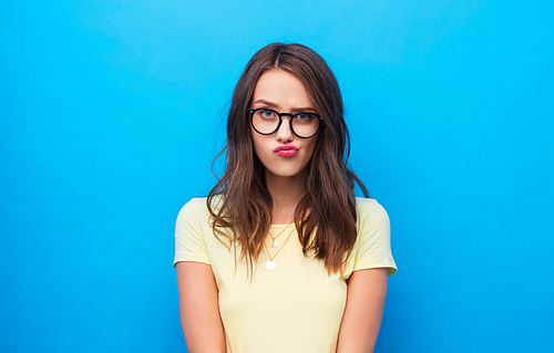 people concept - frowning young woman or teenage girl in yellow t-shirt and glasses over bright blue background