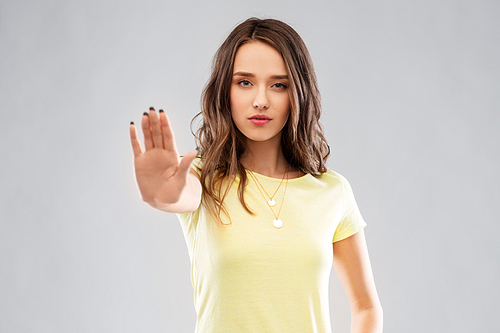 warning and people concept - serious young woman or teenage girl in blank yellow t-shirt showing stop gesture over grey background