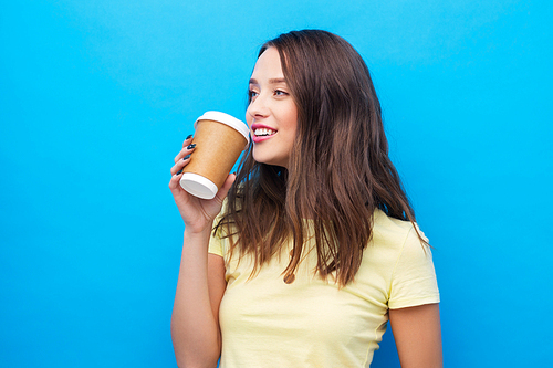 takeaway and people concept - smiling young woman or teenage girl in yellow t-shirt drinking coffee over bright blue background