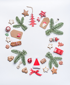 Round Christmas frame layout composition with green fir brunches, craft paper tags, holiday cookies, chocolate, Santa hat, gingerbread and red festive decoration on white background, top view.