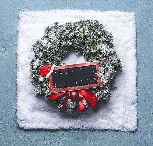 Christmas wreath with green fir branches and red framed sign and Santa hat in snow on blue background, top view with chalkboard copy space for your text or design. Holiday concept. Creative layout