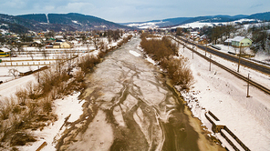 Frozen winter river. Aerial rural view of snowy village, road and railway. Melting snow and ice in early spring.