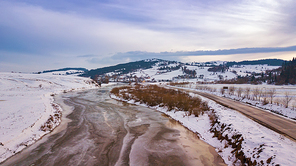Frozen winter river. Aerial rural view of snowy village and road. Melting snow and ice in early spring.