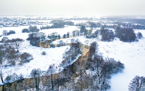 Aerial view of snowy river in winter. Frozen trees on a riverbank and meadow in Belarus near Minsk. Village on background