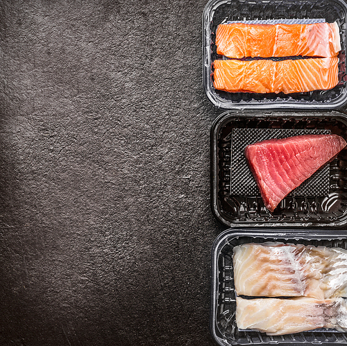 Various of raw fish fillets : salmon, tuna and codfish in plastic boxes on dark rustic background, top view, border, place for text. Healthy Seafood concept