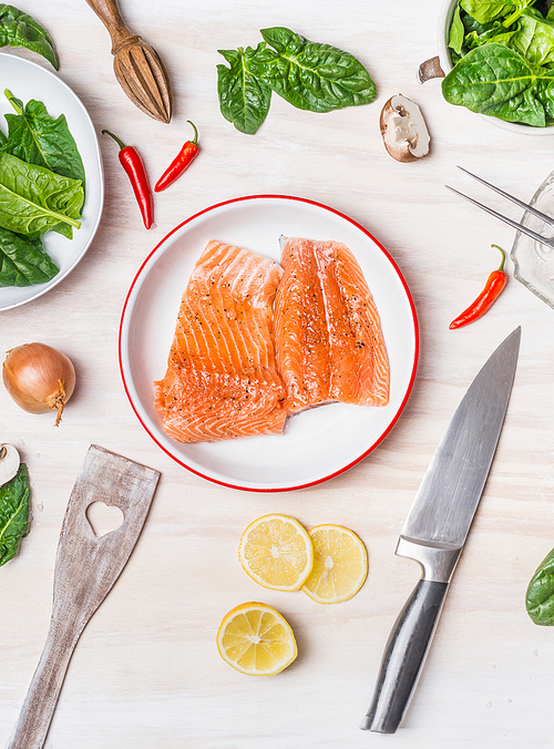 Raw salmon portion in bowl on white kitchen table background with cooking ingredients, spoon and knife, top view
