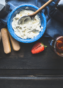 Blue bowl with mascarpone cream and wooden cooking spoon on dark wooden background, top view. Tiramisu preparation