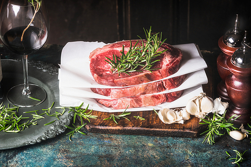 Stack of  raw steaks with rosemary ,spices and grass of red wine on kitchen table at dark wooden background, side view
