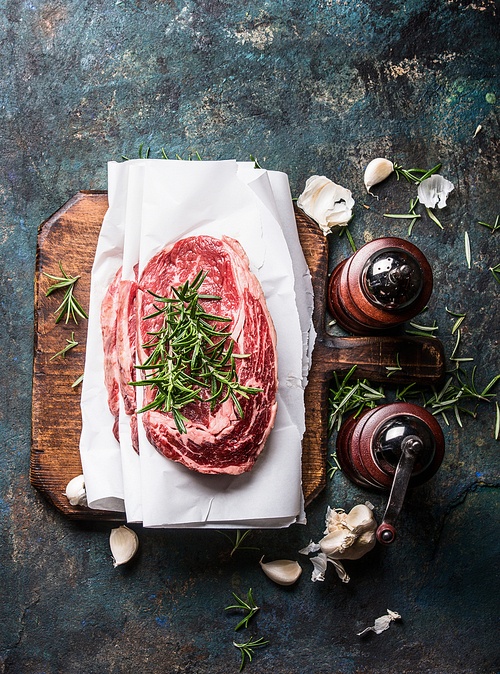 Raw marbled meat steaks with wrapping paper on rustic cutting board with fresh flavoring and spice mills on dark rustic background, top view