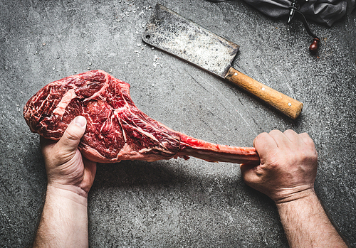 Raw tomahawk beef steak in male hands with meat cleaver on dark rustic background. Butcher hands holding tomahawk steak, top view
