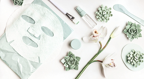 Skin care flat lay with facial sheet mask, mist spray bottle , succulents and orchid flowers on white desktop background, top view. Beauty spa and wellness concept