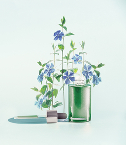Herbal skin care cosmetics and beauty concept. Green facial Serum or oil bottle with dropper or pipette and medical flowers and herbs stand at  blue background, front view.