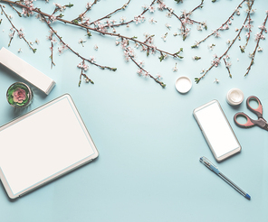 Modern desktop workplace flat lay with mock up of tablet computer and smart phone, cosmetic products, spring blossom branches , scissors, pen  and green leaves on blue background, top view, frame