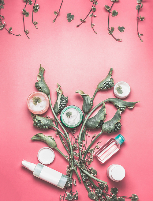 Beauty concept with various facial cosmetic products with mock up, leaves and green flowers on pastel pink desktop background. Modern skin care layout, top view, flat lay.