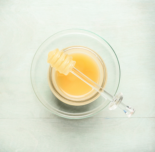 Glass jar with honey and dipper on light blue background, top view