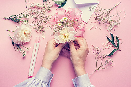 Female hands making lovely floral arrangement with flowers and ribbon on pale pink background, top view. Creative greeting, Invitation and holiday concept