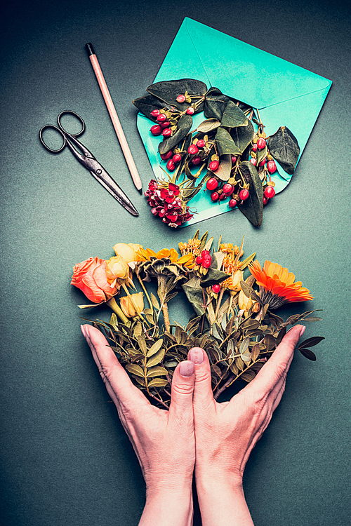 Female hands holding a Creative autumn flowers bouquet on florist workspace table background with opened envelop , shears , pencil, top view.  Invitation , greeting and holiday, concept