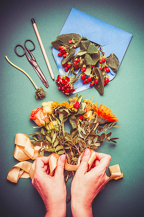 Female hand making autumn flowers arrangements on table workspace with scissors , envelope and ribbon, top view