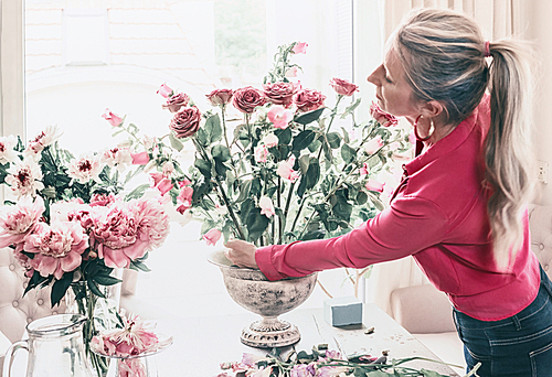 Florist women in red shirt, make beautiful big festive event classical bouquet with roses and other flowers in urn vase on table at window, lifestyle