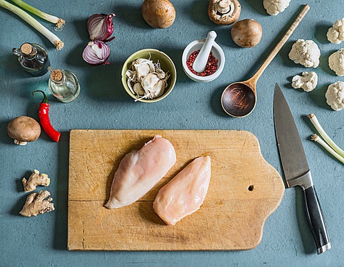Two chicken breast on cutting board with knife on kitchen table background with ingredients , top view. Cooking preparation