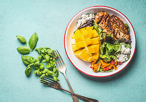 Tasty summer salad with roasted chicken breast and mango in bowl with cutlery on light blue background, top view. Healthy low carb lunch