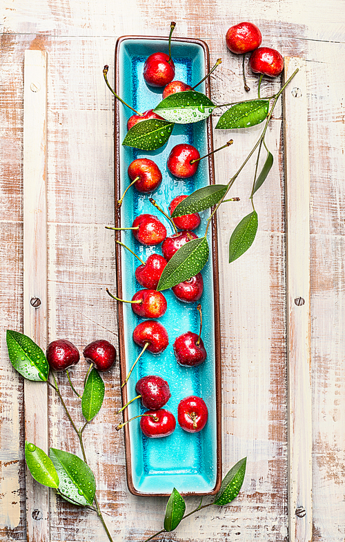 Sweet cherries with green leaves on blue rectangular plate on light wooden rustic background, top view. Summer fruits and berries concept.