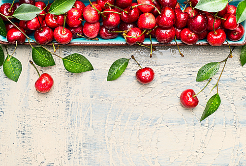 Border of sweet cherries with green leaves on light wooden background, top view, place for text. Summer fruits and berries concept.