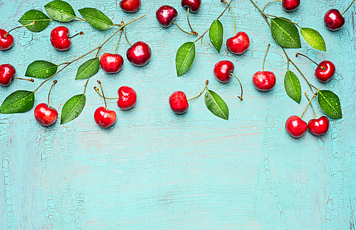 Border of sweet cherries on branch with green leaves on light blue background, top view, place for text. Summer fruits and berries concept.