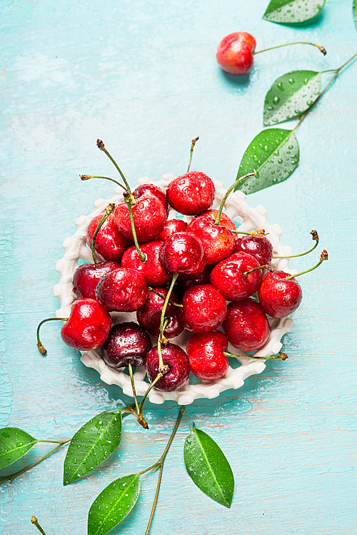 Ripe sweet cherry with leaves in white bowl on blue wooden background, close up