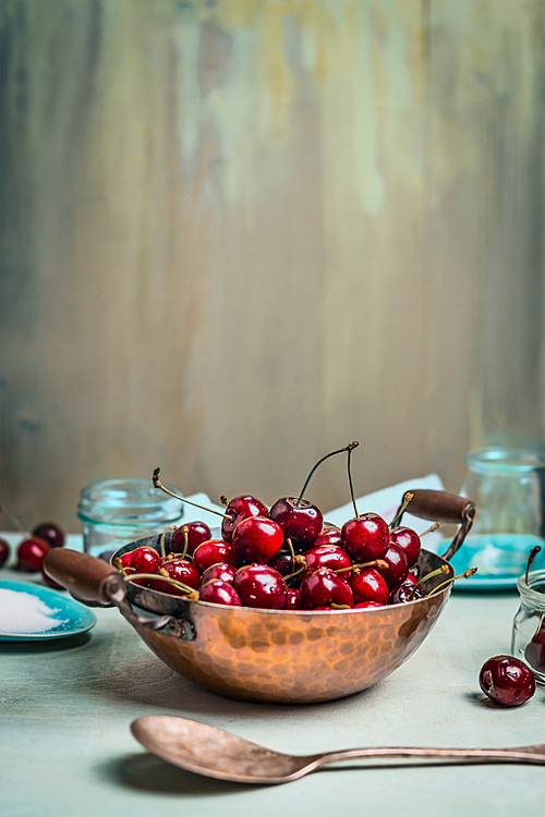 Sweet cherries in old copper pot with glass jars and spoon on light  blue rustic wooden background, place for text