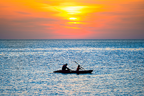 Orange sunset over blue sea and silhouette of kayak in rippled water