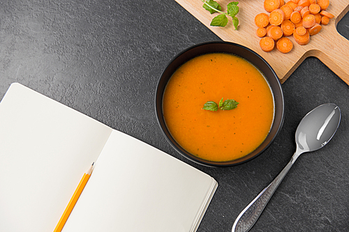 food, new nordic cuisine, culinary and cooking concept - close up of vegetable pumpkin cream soup in bowl, spoon and notebook with pencil on stone table