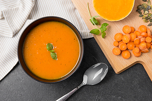 food, new nordic cuisine, culinary and cooking concept - close up of pumpkin cream soup in bowl, spoon and cutting board with vegetables on slate stone background