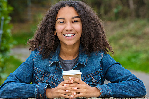 Beautiful happy biracial mixed race African American girl teenager female young woman smiling with perfect teeth drinking takeaway coffee outside wearing a denim jacket and leaning on a wall