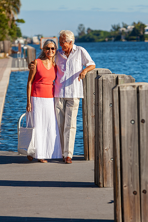 Happy wealthy successful senior man and woman romantic couple together walking and embracing by tropical sea or river with bright clear blue sky