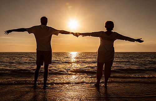 Senior man and woman couple holding hands arms wide at sunset or sunrise on a deserted tropical beach