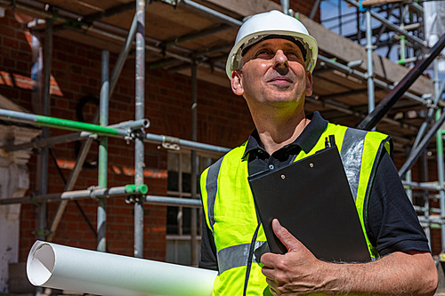 Thoughtful middle aged man, male builder foreman, worker, contractor or architect on construction site holding black clipboard and architectural plans