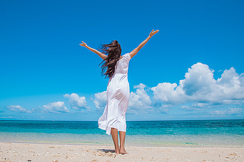 Woman in white dress posing in tropical sea beach with arms raised