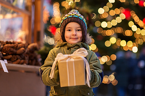 holidays, childhood and people concept - happy little boy with gift box at christmas market in winter evening