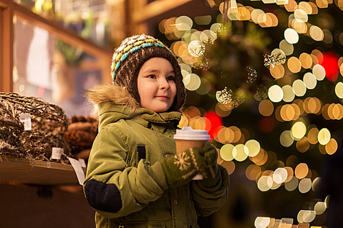 holidays, childhood and people concept - happy little boy with cup of tea at christmas market in winter evening