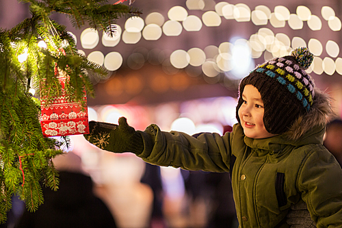 holidays, childhood and people concept - happy little boy reaching to gift bag hanging on fir tree at christmas market in evening