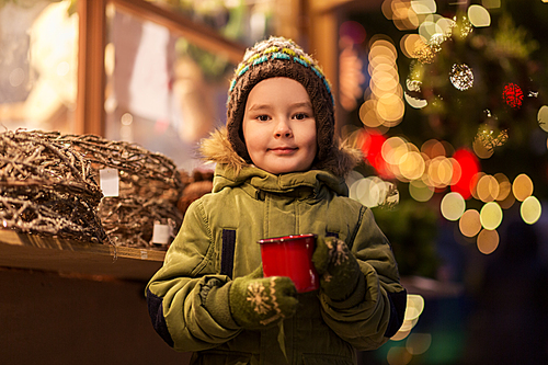 holidays, childhood and people concept - happy little boy with cup of tea at christmas market in winter evening
