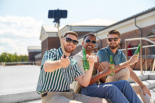 leisure, technology and people concept - happy male friends taking picture by smartphone on selfie stick and drinking beer on street in summer
