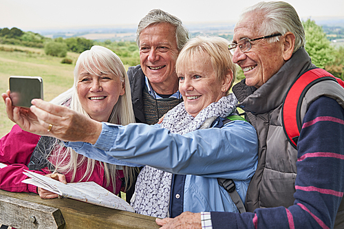 Group Of Senior Friends Hiking In Countryside Standing By Gate  And Taking Selfie On Mobile Phone