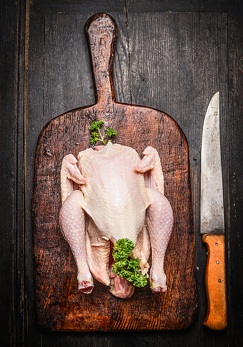 Raw whole chicken on vintage cutting board with kitchen knife on rustic wooden background, top view